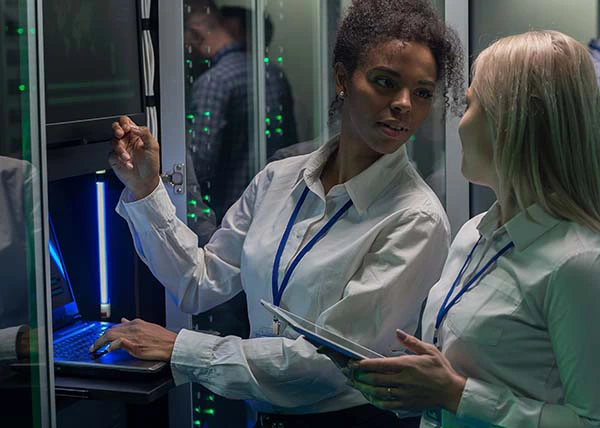 Two women in white shirts and ID lanyards discussing work near servers and a laptop in a data center.