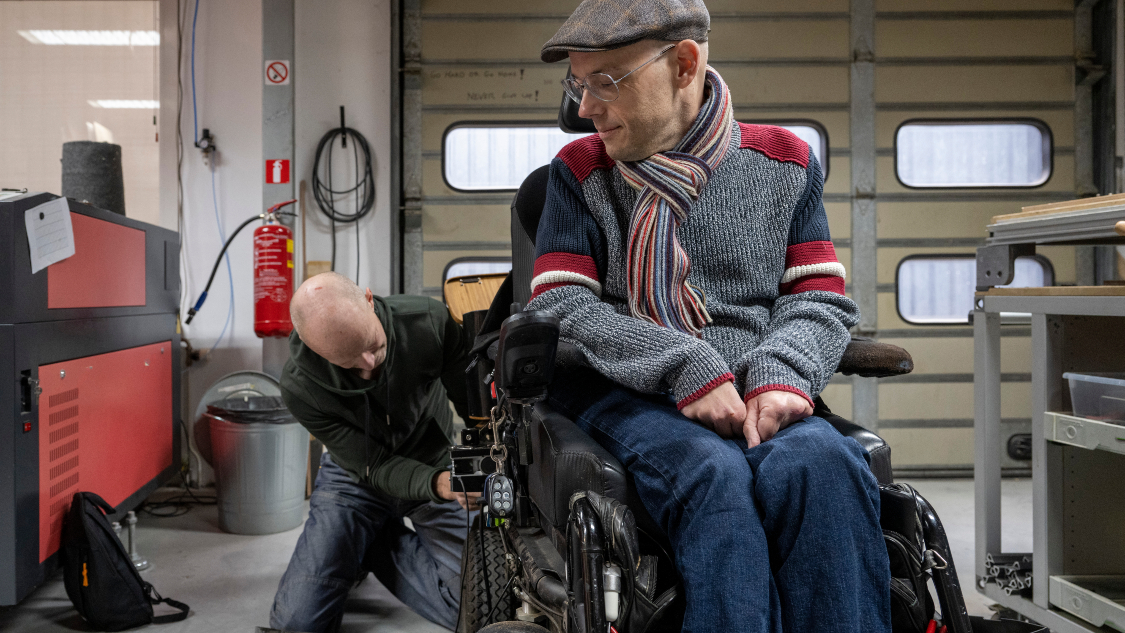 In a workshop setting, a technician kneels beside a man in a powered wheelchair, performing maintenance or adjustments. The man wears a cap, glasses, and a striped scarf, and observes attentively. Tools and machinery are visible in the background.