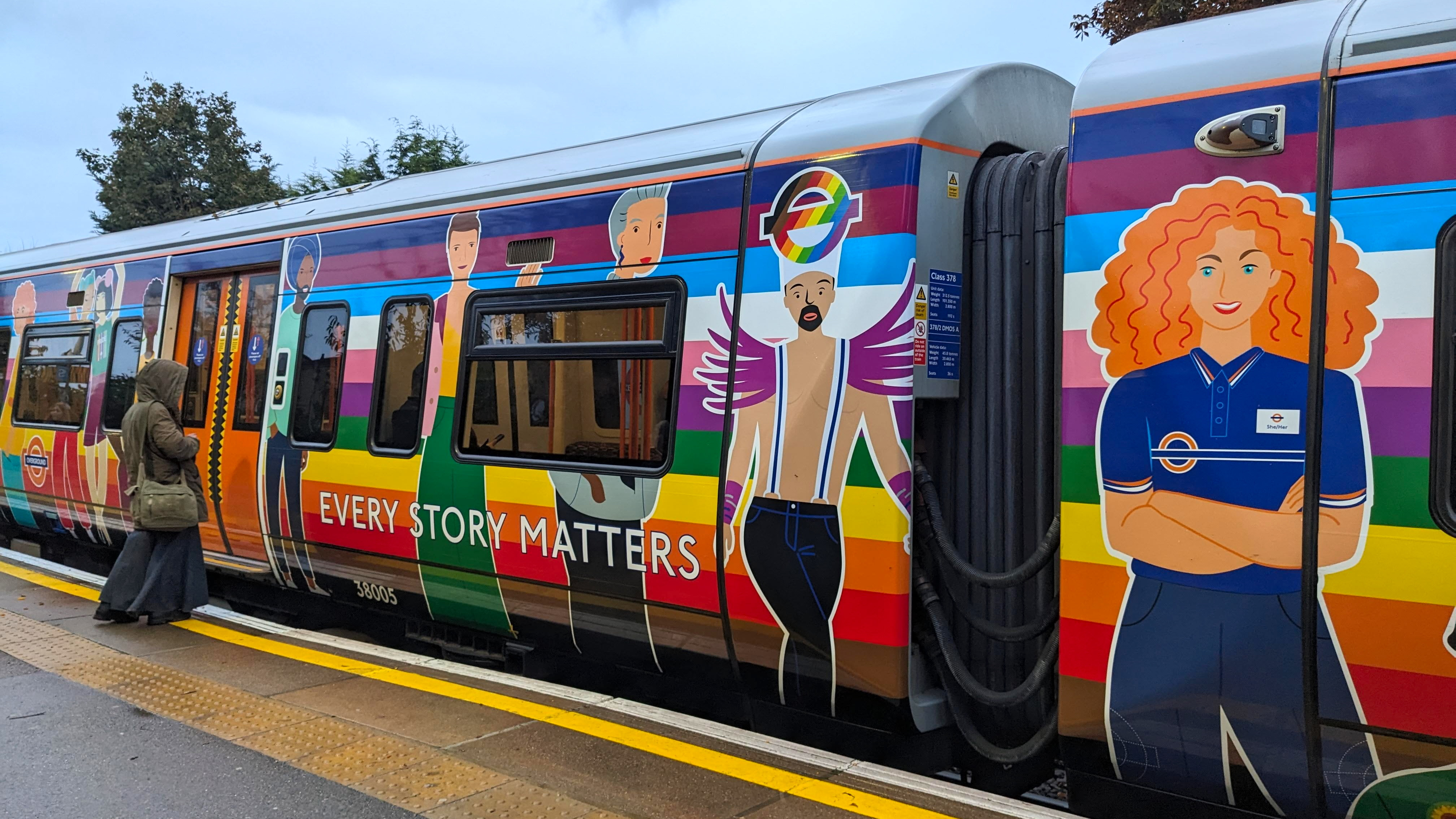A London Overground train decorated with rainbow colours and cartoon illustrations of diverse people, featuring the slogan “EVERY STORY MATTERS.” A person stands on the platform, looking toward the train.