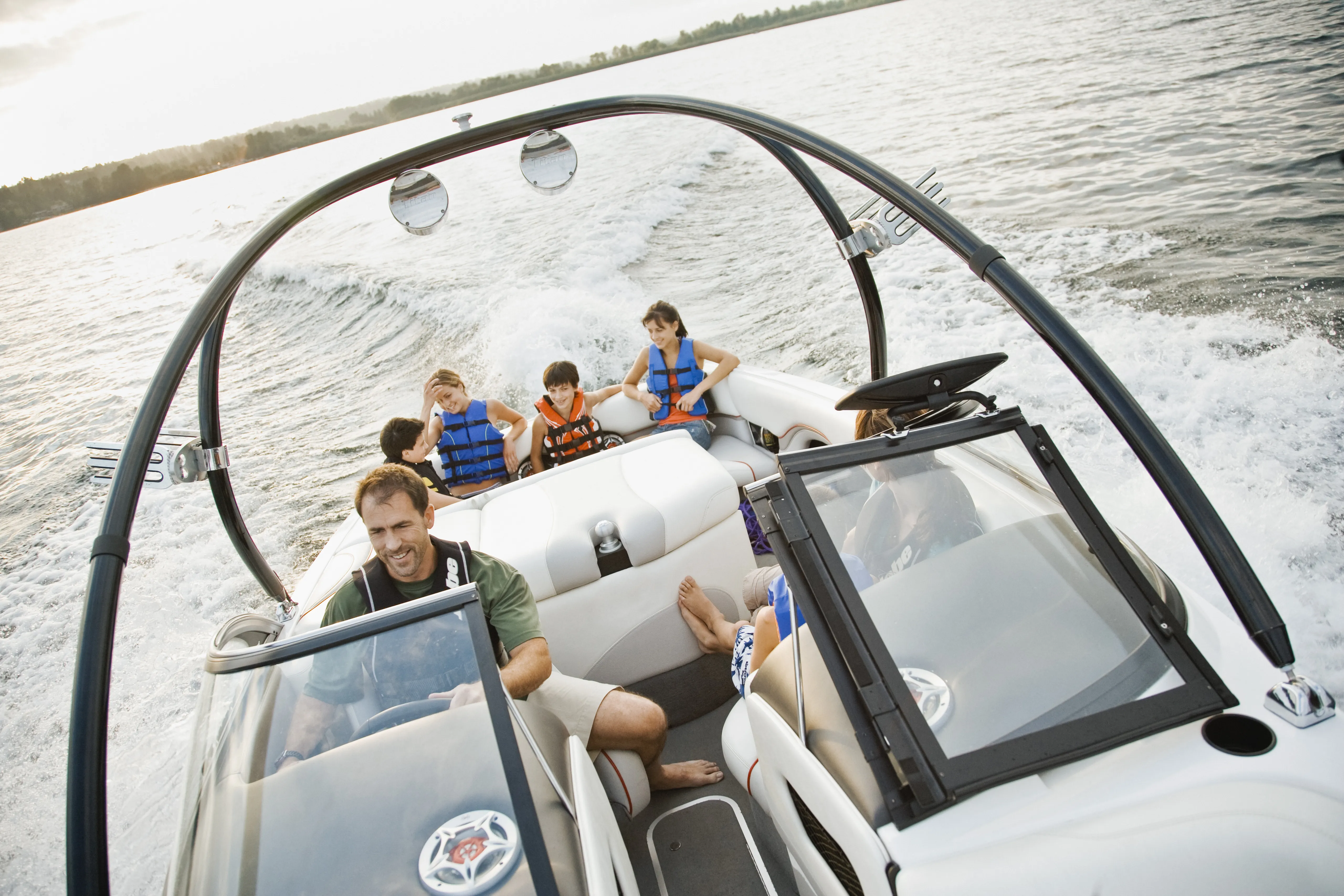 A family wearing life jackets, sit in a moving speedboat on a lake. The boat leaves a foamy wake, with water and a distant tree-lined shoreline visible in the background. 