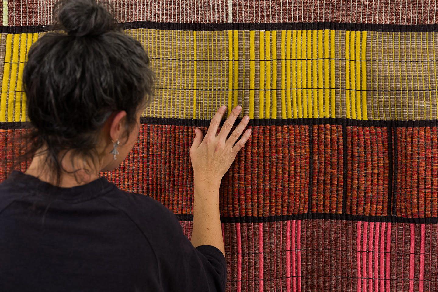 Artist Rachel Dana, wearing black, touches a yellow and grey section of her woven art.