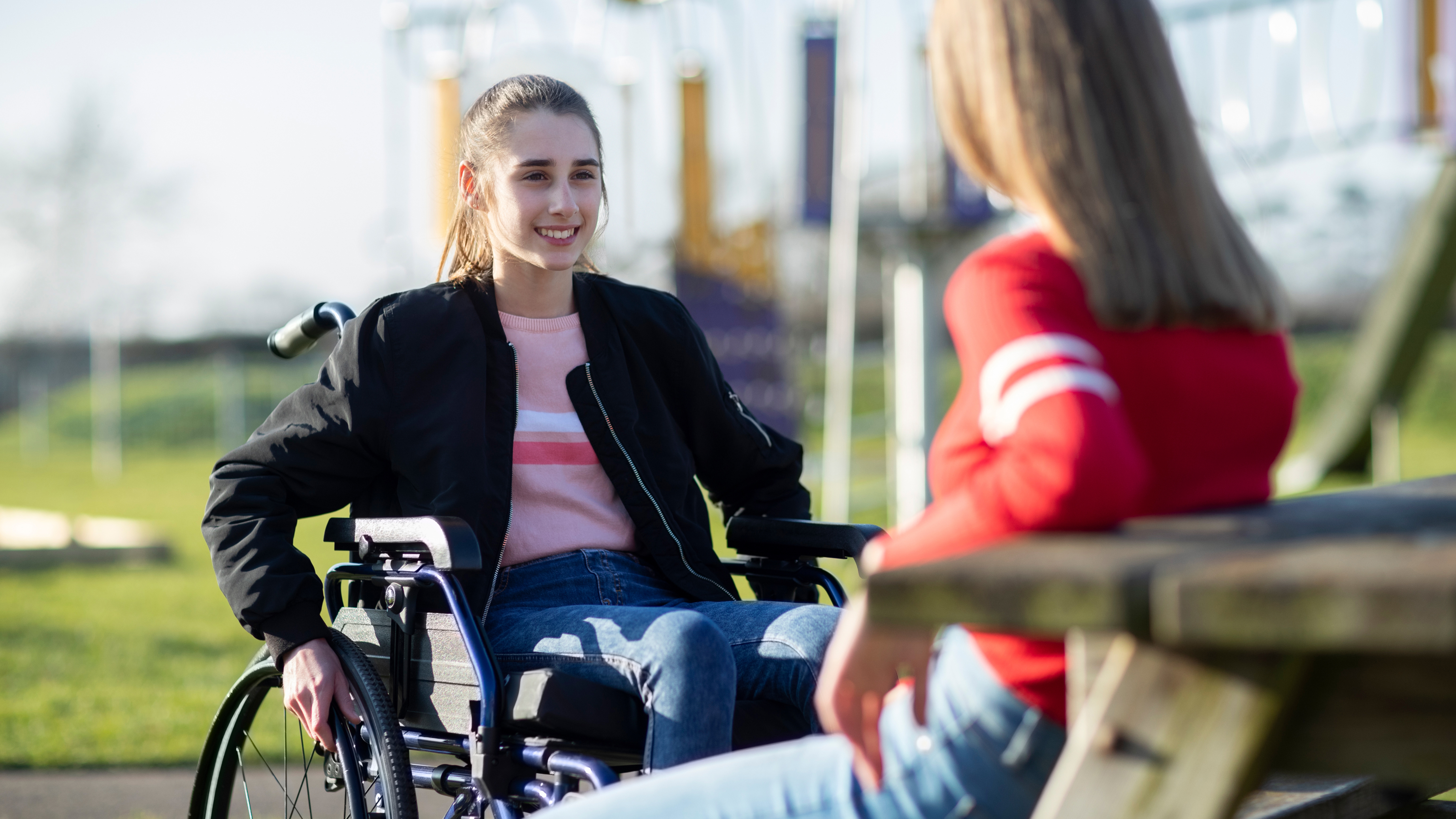 A girl in a wheelchair smiles while talking with a friend who is seated at a wooden picnic table in a sunny outdoor park. The background features green grass, play equipment, and soft natural lighting.