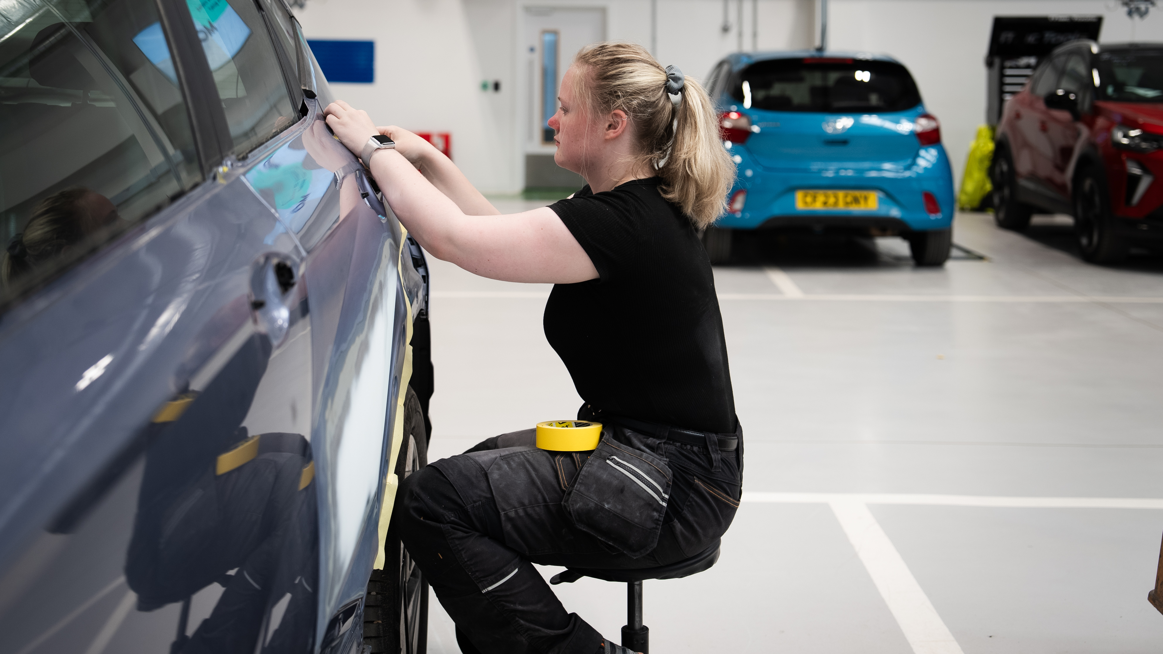 A woman in a black shirt and work trousers sits on a stool while preparing the side of a car for bodywork or paint repair inside a vehicle workshop. A roll of masking tape rests on her lap, and other cars are parked in the background.