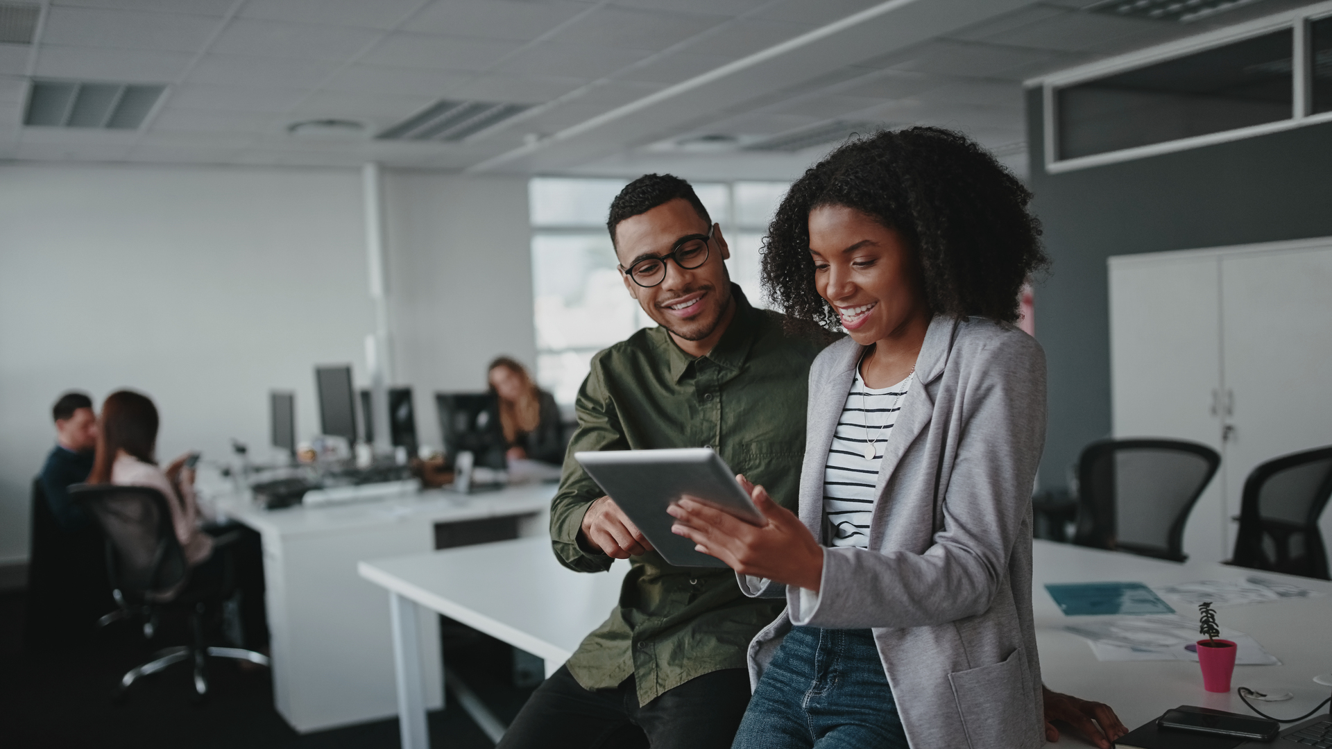 Successful two african american young businesspeople sitting on desk using digital tablet while colleague in background at office