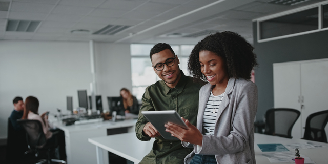 Successful two african american young businesspeople sitting on desk using digital tablet while colleague in background at office