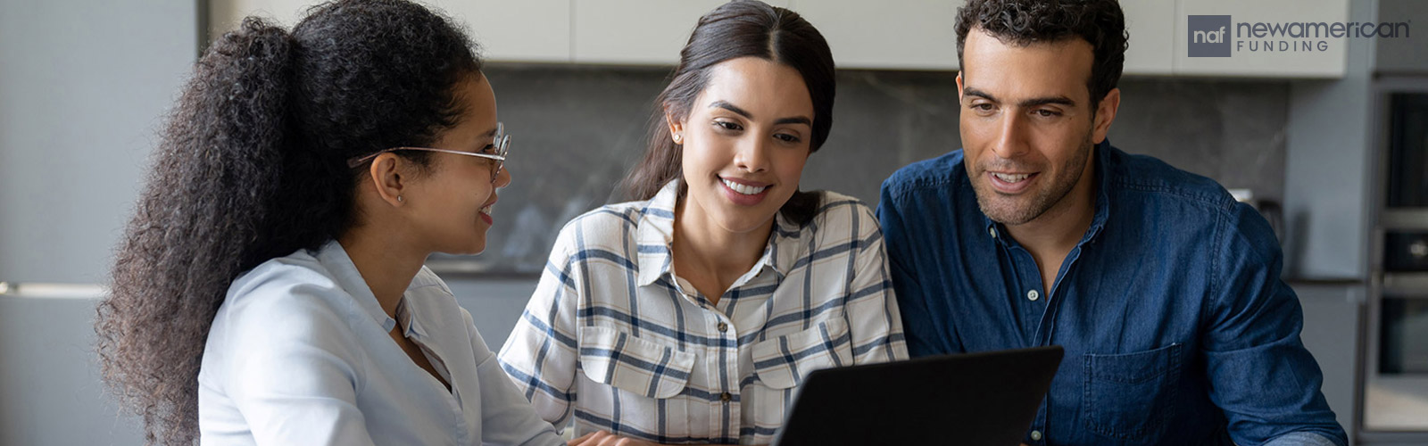 A woman sits at a table with another woman and man, she is showing them something on a laptop and they are all smiling