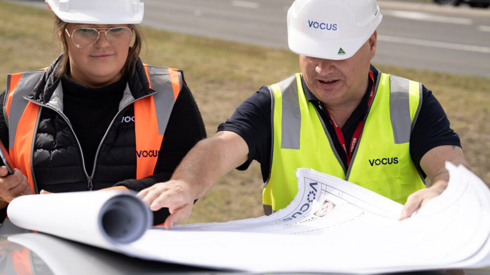 Image of two Vocus workers with hard hats on unrolling plans. They're both wearing high vis shirts