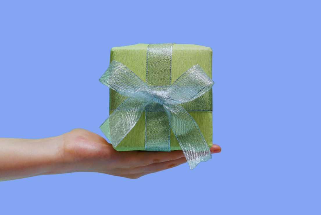 Hand holding out a present wrapped in green wrapping paper and ribbon against a blue background.