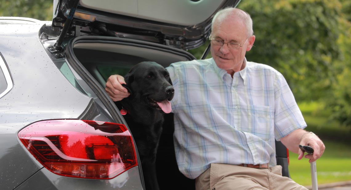 An older man with glasses and a walking stick sits at the open boot of a car, petting a black Labrador dog inside the vehicle.