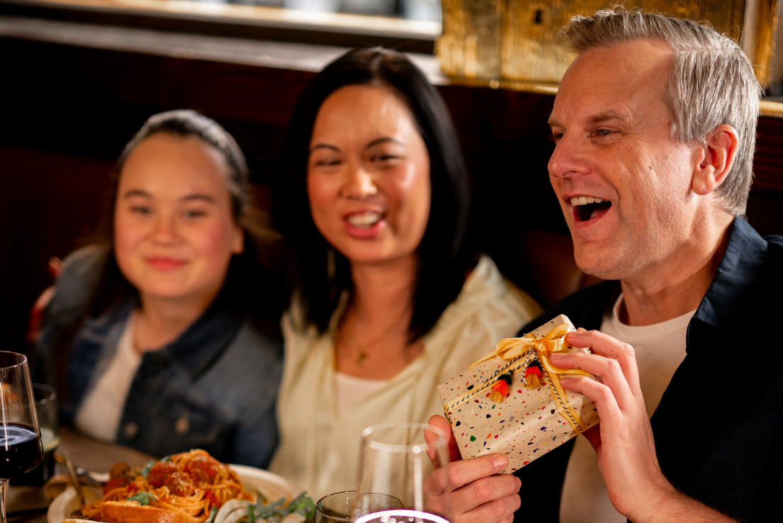 Dad holding a wrapped gift during a Father’s Day family meal at a restaurant with his wife and daughter.