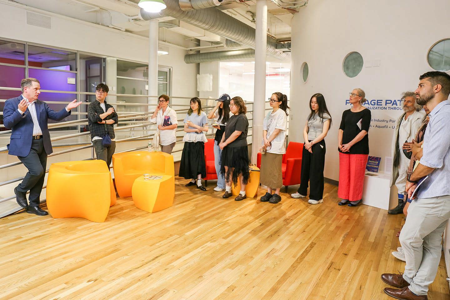 Ian Burt stands in a large room, speaking to a group of MFA Textiles students and faculty standing before him.