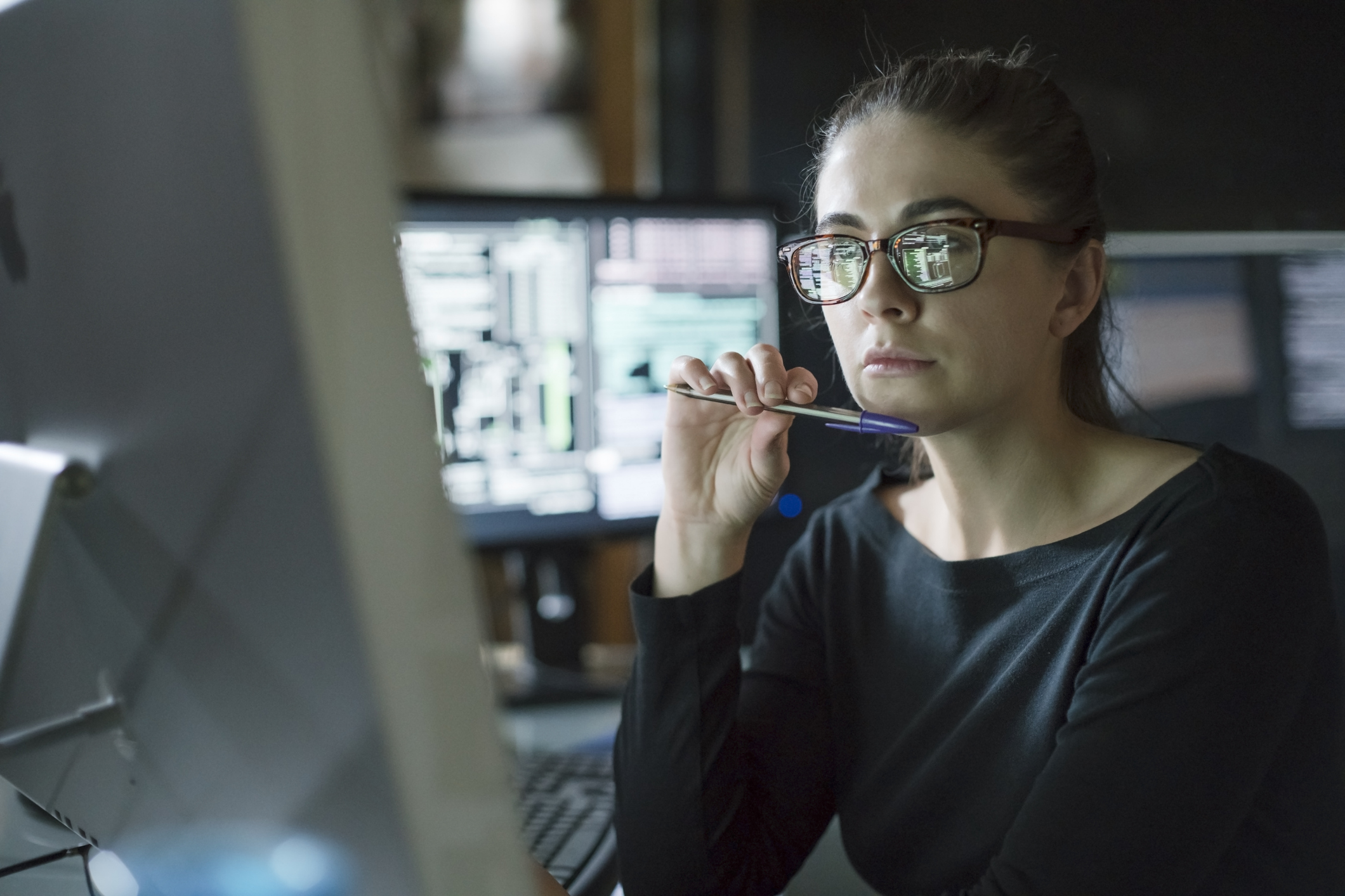 A software engineer with glasses works late in a dark office, her face illuminated by the code reflected from her computer screen.
