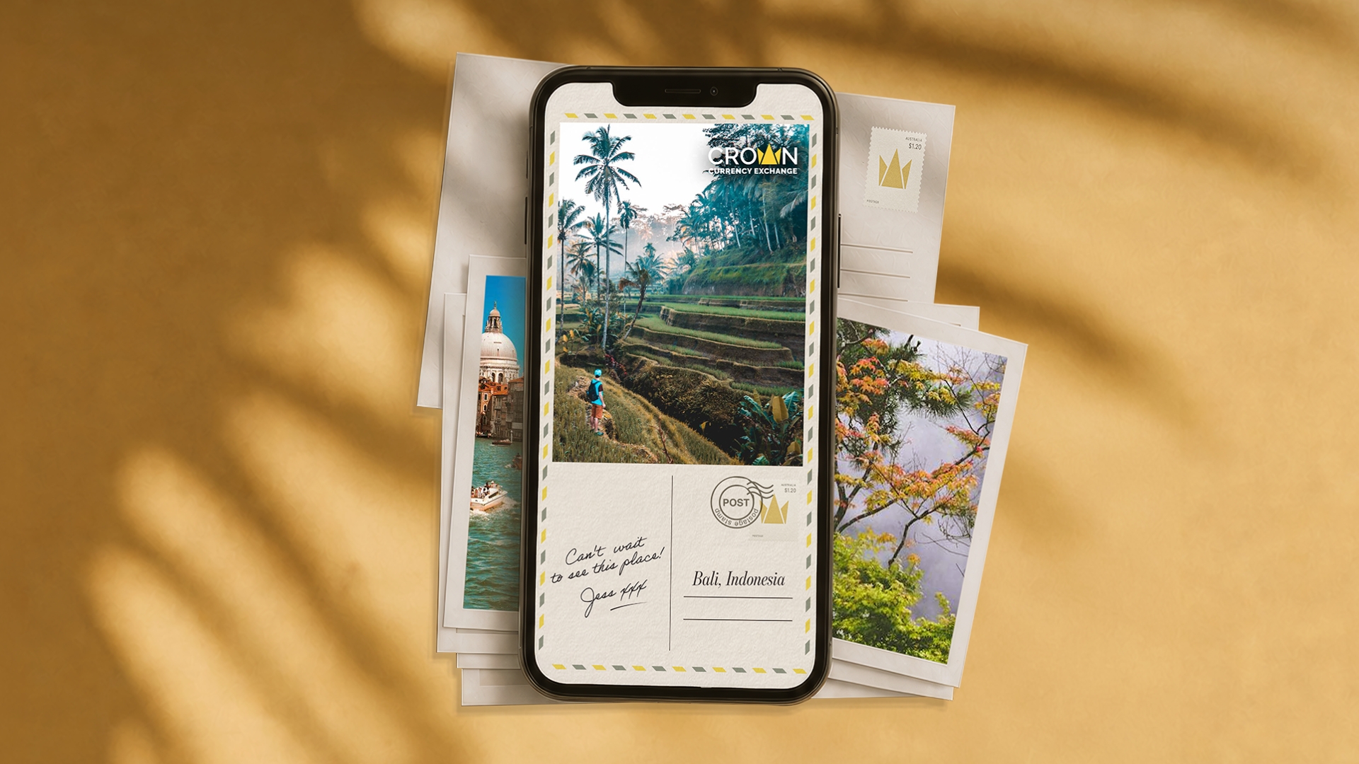 Smartphone displaying a Crown Currency Exchange postcard ad styled as a vintage airmail postcard, showing a Bali rice terrace scene with a hiker, a handwritten note reading “Can't wait to see this place! Jess xxx,” and the caption “Bali, Indonesia.” Travel photo prints of a European canal and autumn foliage are scattered behind it on a warm golden background.