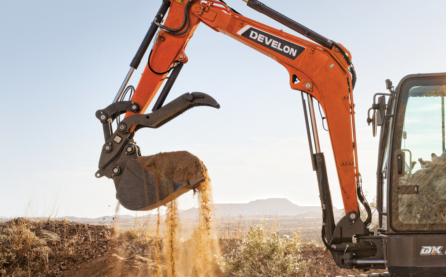 A DEVELON mini excavator scooping a bucket of dirt on a stretch of open farmland.