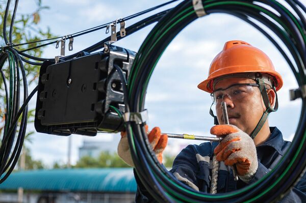 Telecommunications technician wearing a hard hat and safety glasses works with fiber optic cables and outdoor equipment.