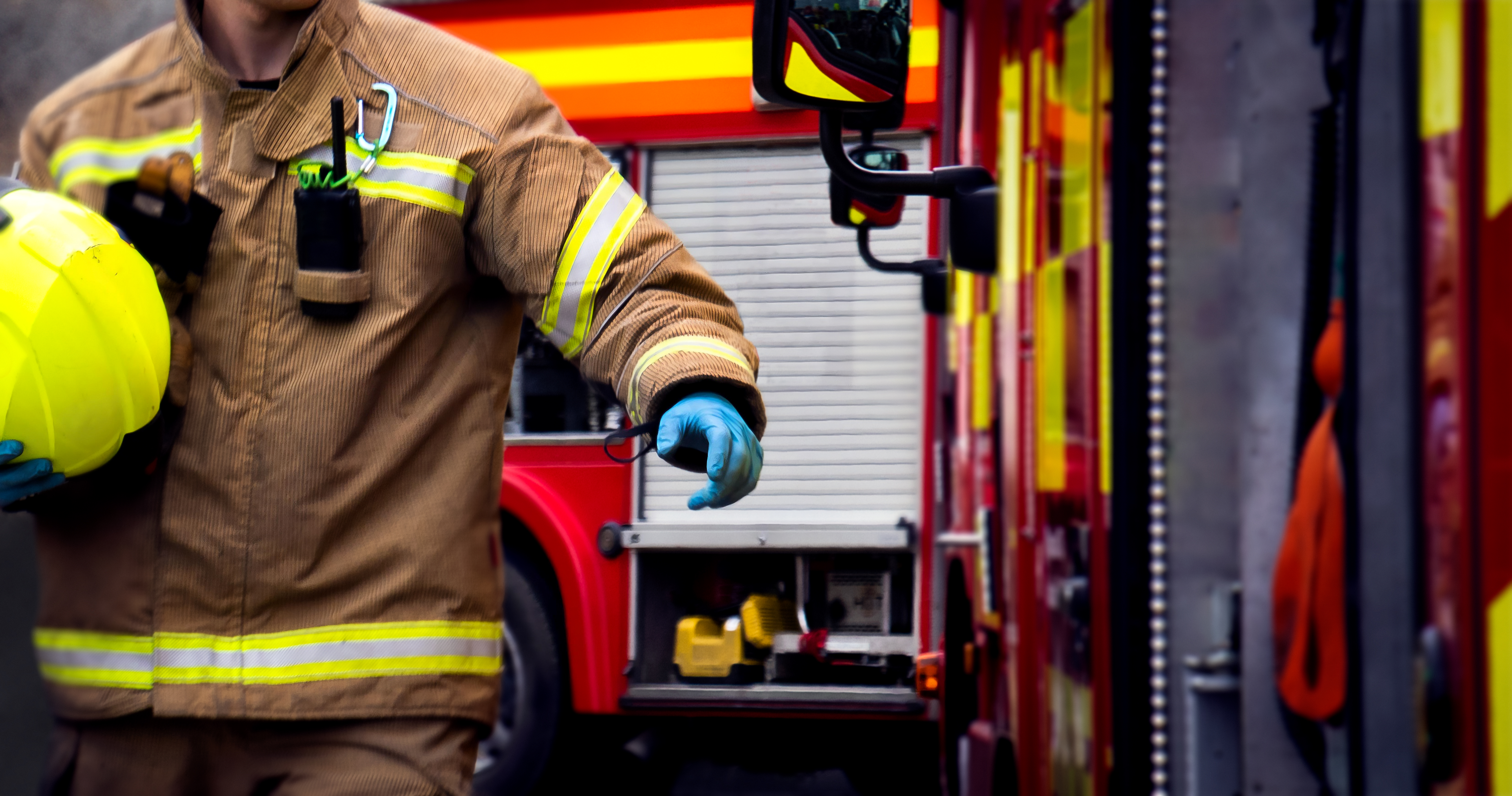 A fire EMS technician approaches a fire response engine wearing firefighter gear and holding a helmet in the fire station. His face is out of frame.