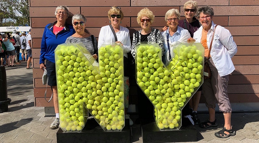 The women stand behind a statue made of tennis balls that forms the shapes of the letters "NY"