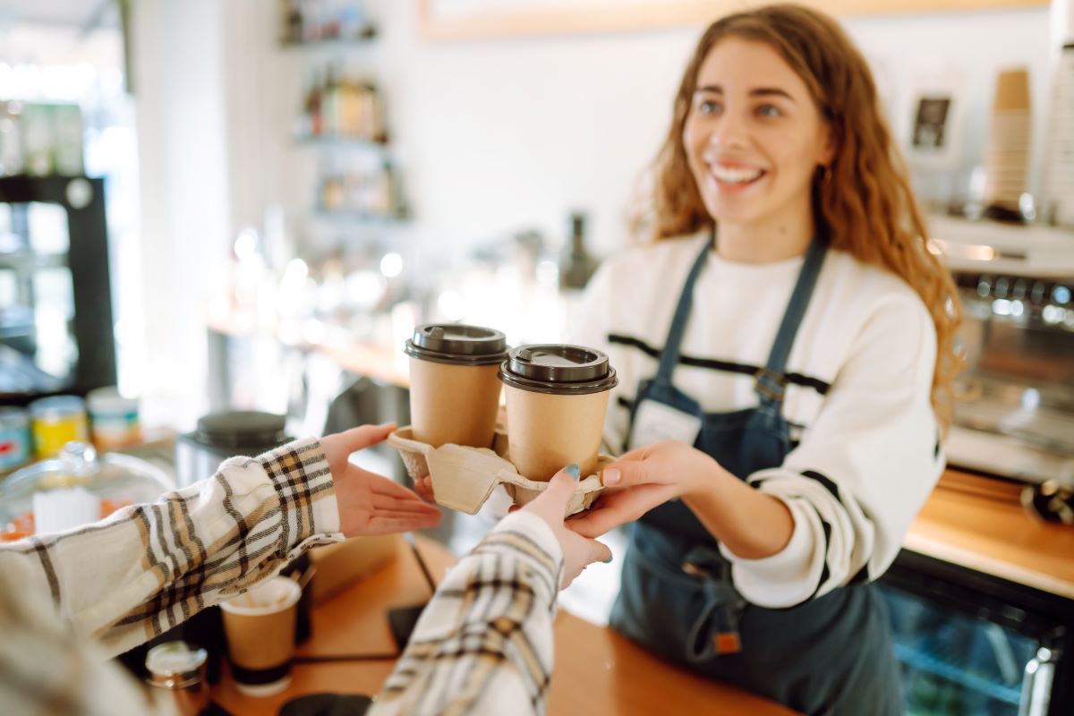 A smiling female barista hands two warm takeout coffee cups to a waiting customer.
