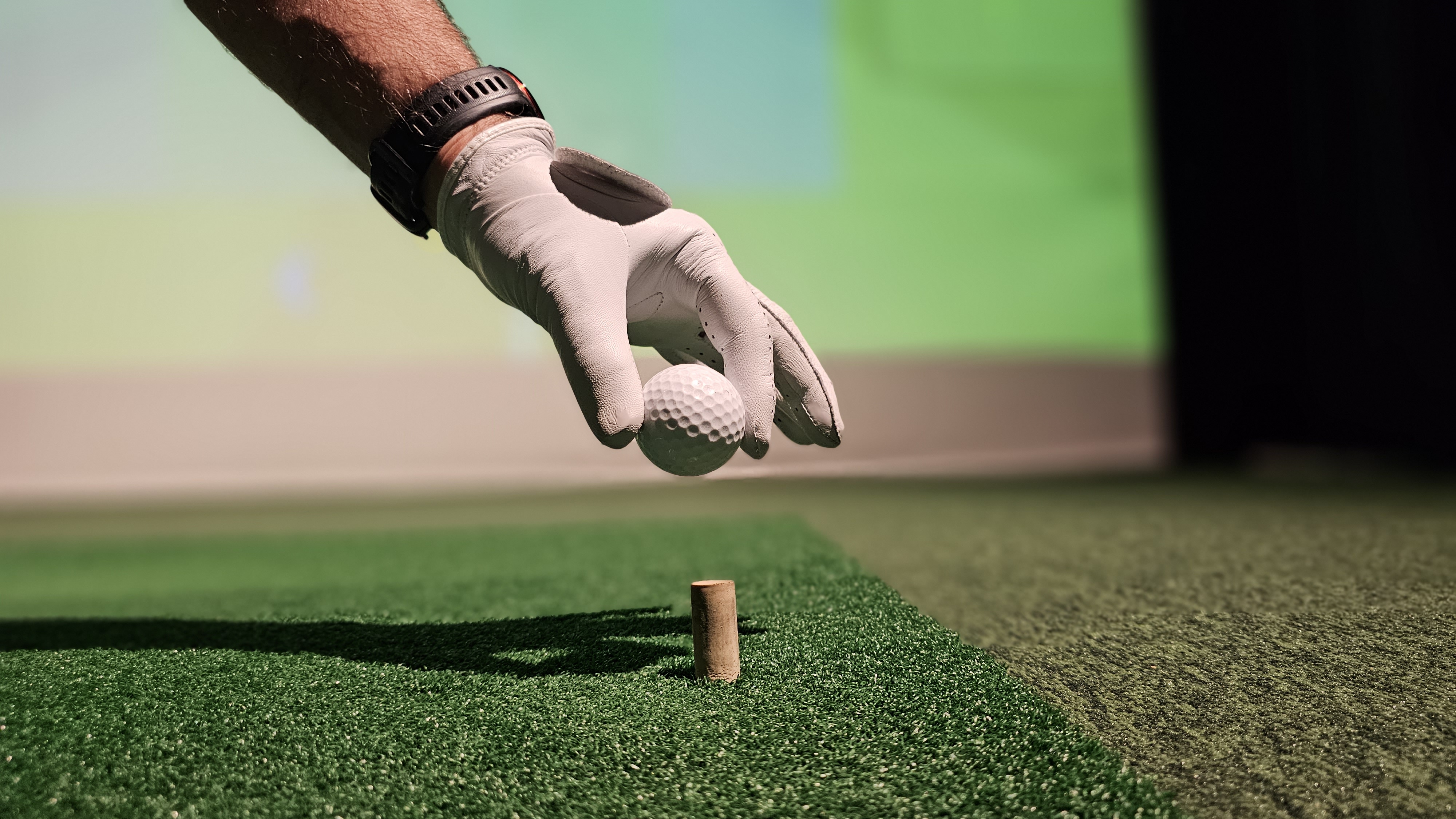 A gloved hand places a golf ball on a putting tee in a sports simulator, using projection simulation technology to capture the nuances of a real game.