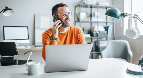 Man on his phone while sitting at a desk on his laptop