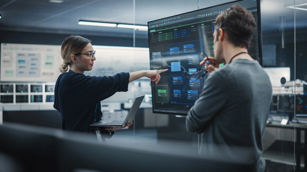Stock image of two workers in an office, pointing at a Desktop screen with data on display