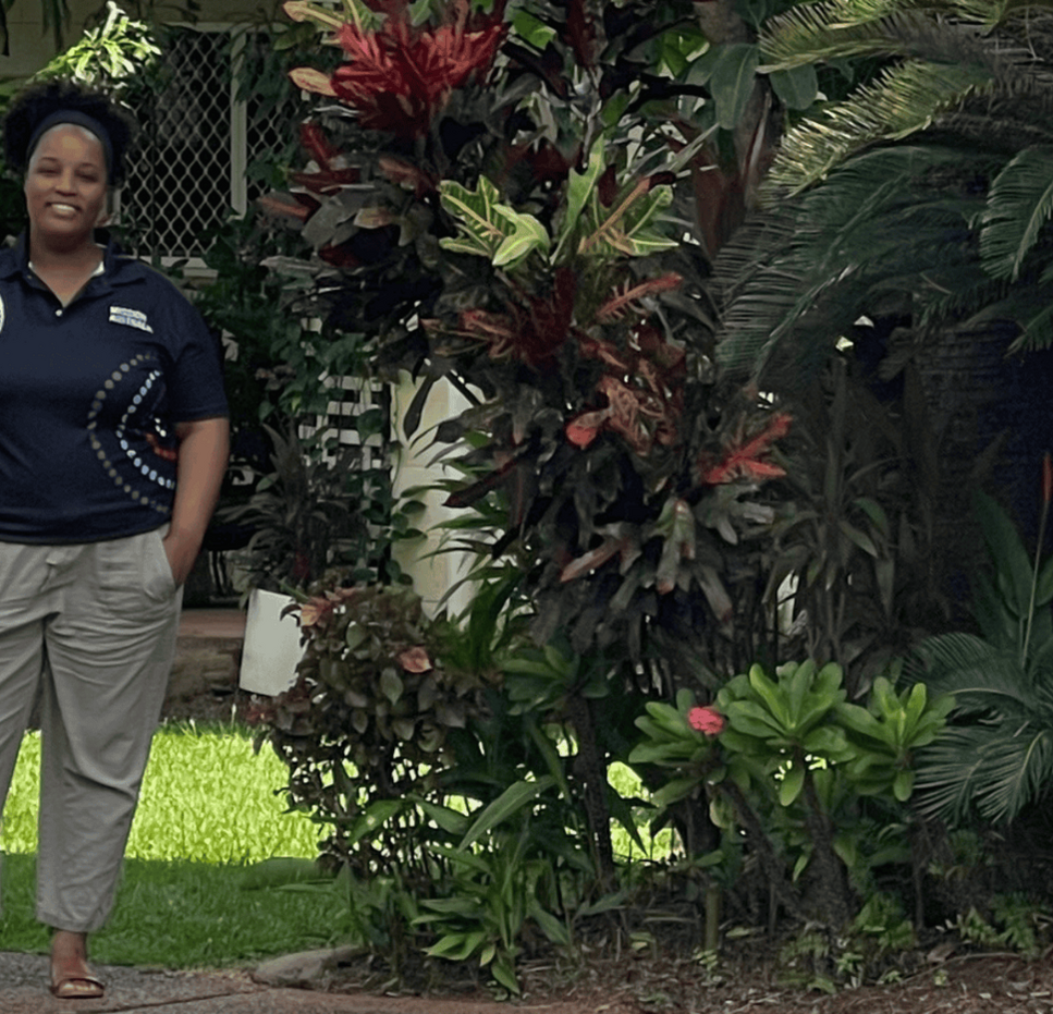 A person standing on a sidewalk in front of a house surrounded by lush greenery and tropical plants.