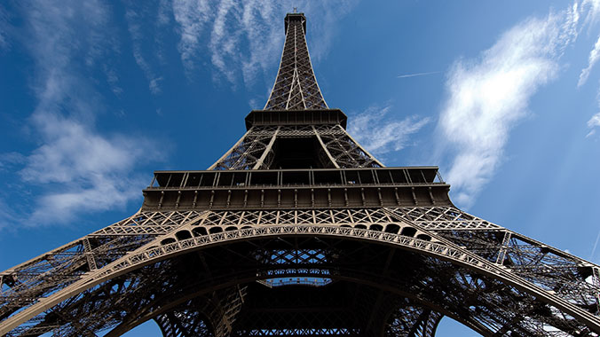 A low-angle view looking up at the iron lattice of the Eiffel Tower in Paris against a blue sky with white clouds.