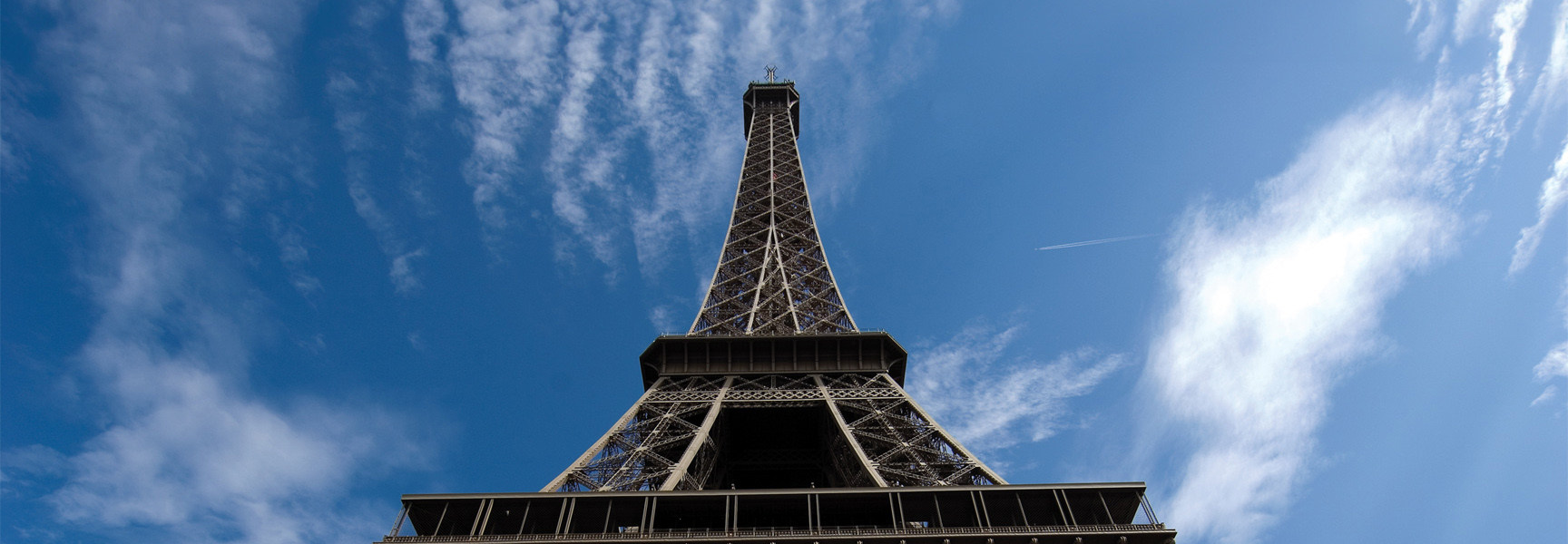 A low-angle view of the Eiffel Tower in Paris, France, stretching up towards a bright blue sky with wispy white clouds.