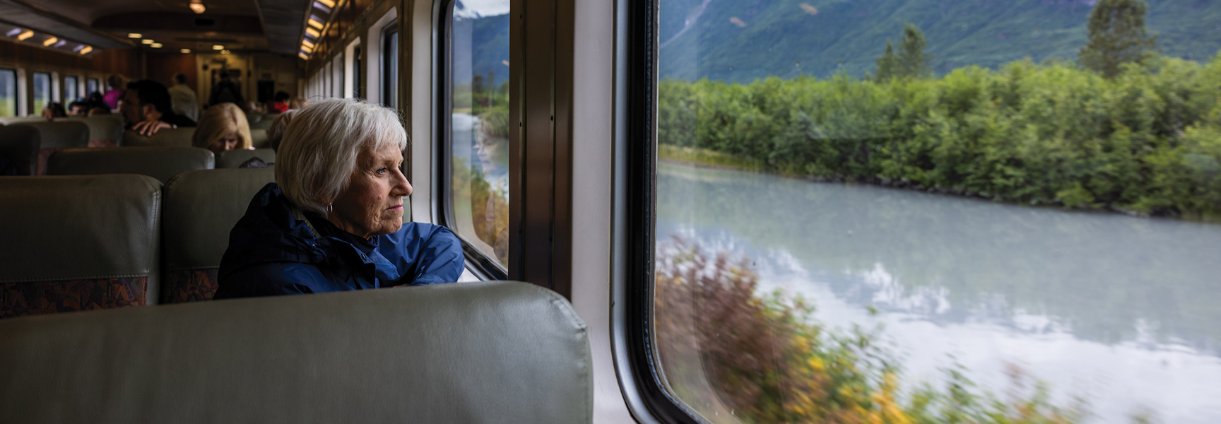 An older woman with white hair looks out the window of a train at the Alaskan scenery passing by.
