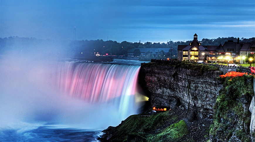 Illuminated Horseshoe Falls at Niagara Falls with the Table Rock Centre building overlooking the rushing water at dusk in Ontario.