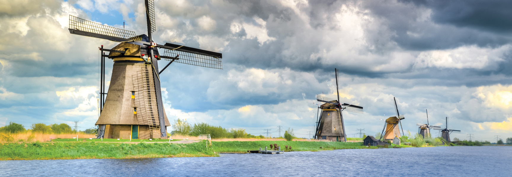 A row of traditional Dutch windmills lines a canal under a dramatic cloudy sky in the Netherlands.