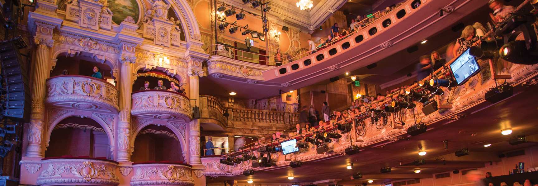 The ornate interior of a classic London theatre, with patrons sitting in lavishly decorated balconies and seating areas before a performance.