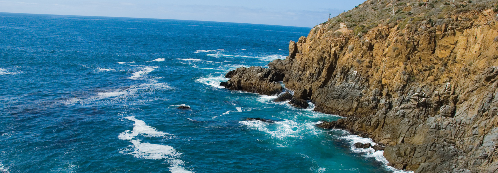 The brilliant blue ocean crashes against the rugged, rocky coastline of Baja, Mexico under a sunny sky.