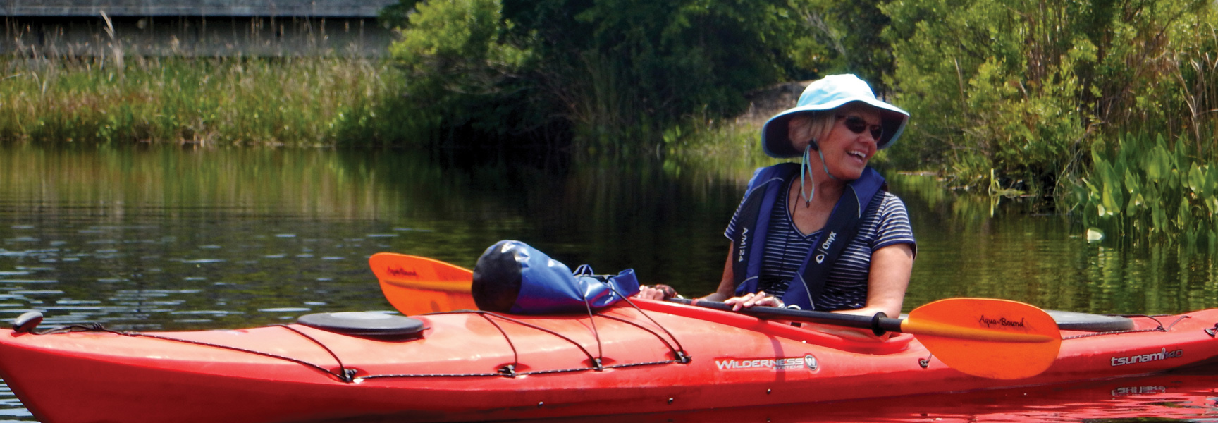 A woman smiles while kayaking on a waterway in South Carolina, surrounded by lush green foliage under a clear sky.