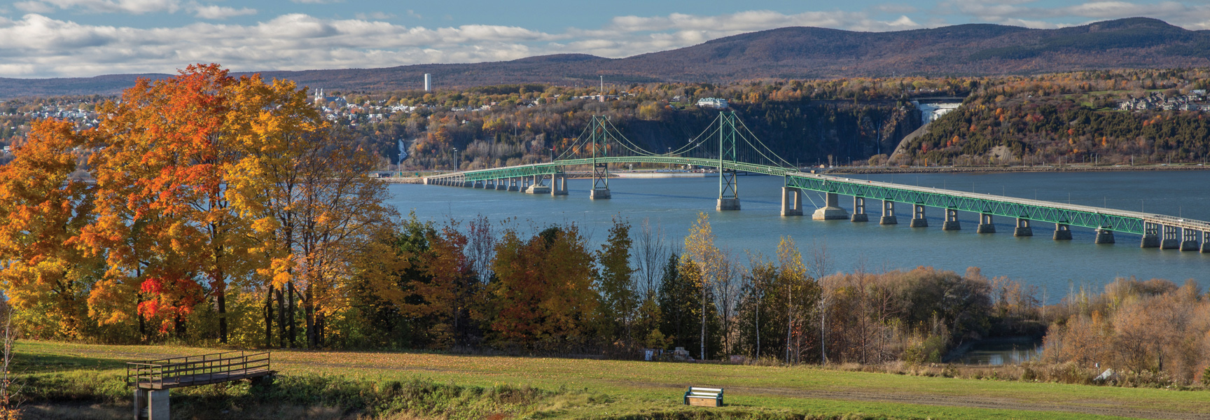 The green Île d'Orléans bridge spans the St. Lawrence River in Québec, framed by vibrant autumn trees and with Montmorency Falls in the distance.