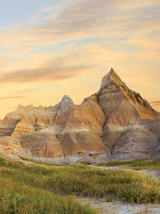 Jagged rock formations in the Badlands of South Dakota stand over a grassy landscape under a soft sunset sky.