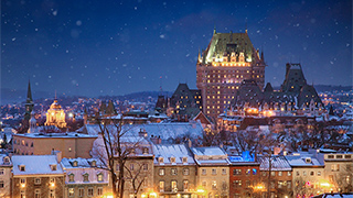 Snow falls at night over the illuminated skyline of Québec City, with the Château Frontenac rising above the snow-covered rooftops.