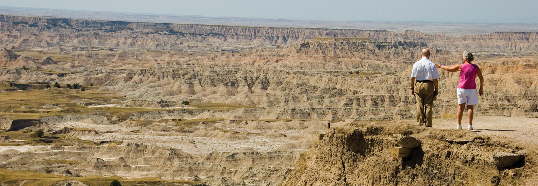 An older couple stands on a cliff's edge, overlooking the expansive, layered rock formations of the Badlands in South Dakota.