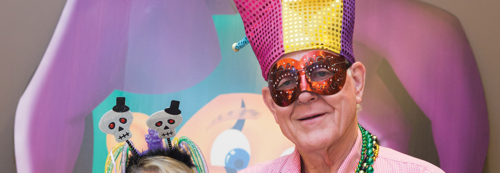 A smiling man wears a colorful jester hat, mask, and beads during Mardi Gras celebrations in New Orleans, Louisiana.