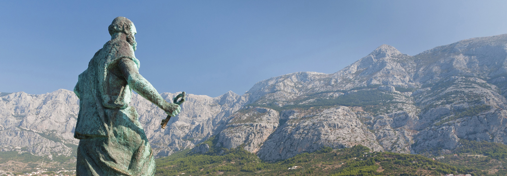 A weathered statue overlooks a vast mountain range rising above the scenic coastline of Croatia.