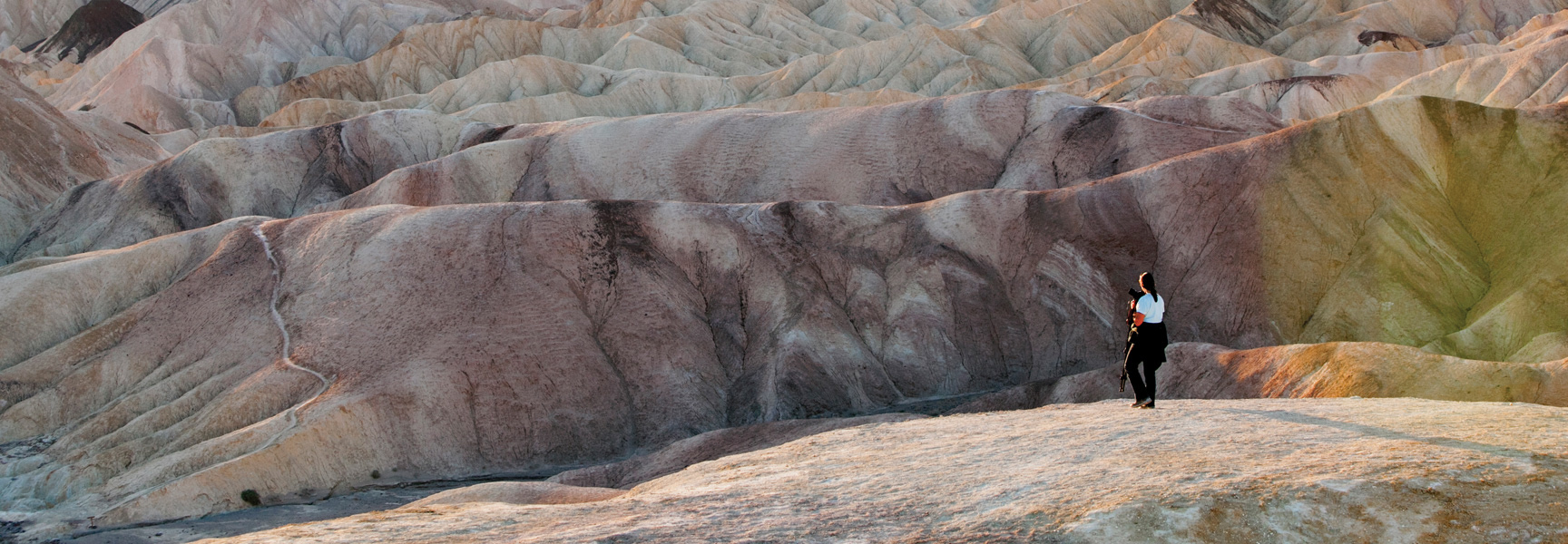 A photographer stands on a ridge overlooking the vast, colorful, and eroded badlands of a desert in Arizona/California at sunset.
