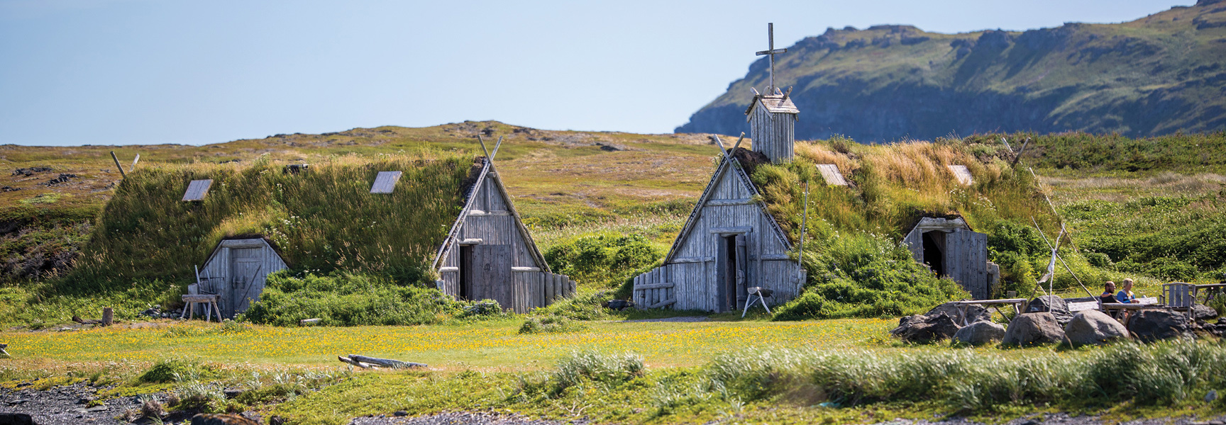Reconstructed Viking sod-roof houses sit in a grassy field with rolling green hills behind them in Newfoundland and Labrador.