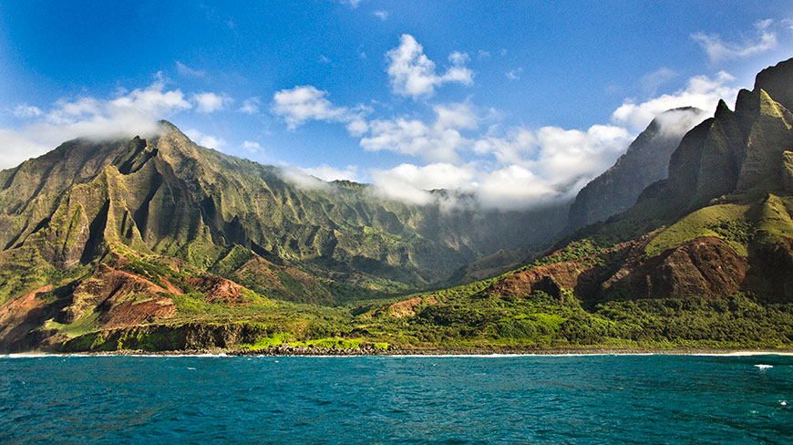 The dramatic, cloud-covered green cliffs of Hawaii's Waimea Canyon rise steeply from the deep blue ocean under a partly cloudy sky.