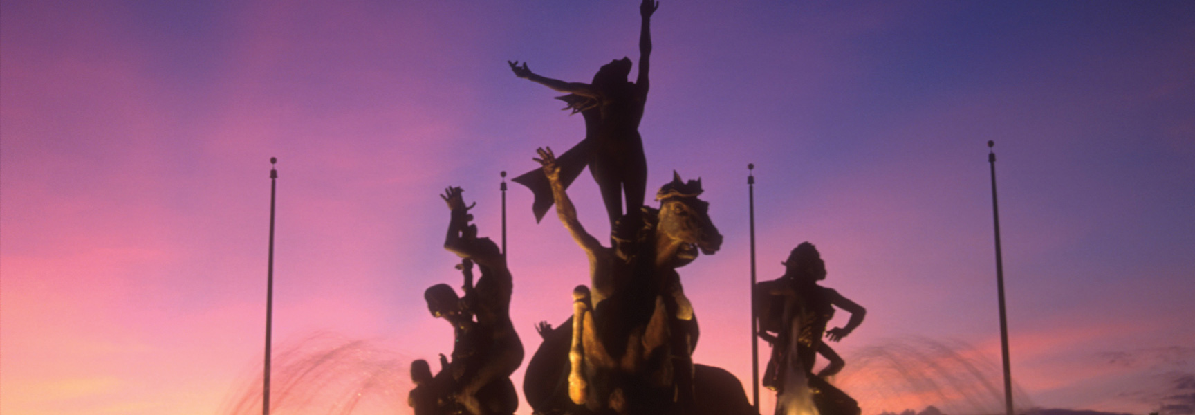 The silhouette of a historic fountain statue in Puerto Rico stands against a vibrant purple and pink sunset.