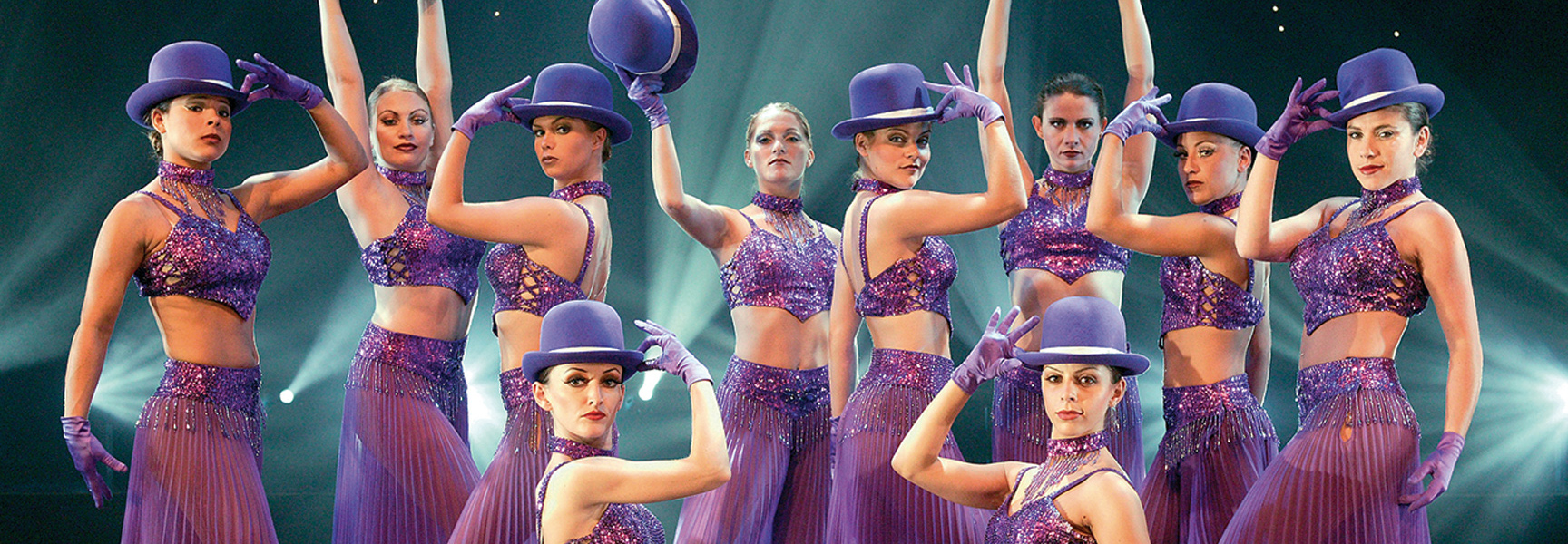 A group of female performers in sparkling purple costumes and bowler hats pose on a stage in Missouri.