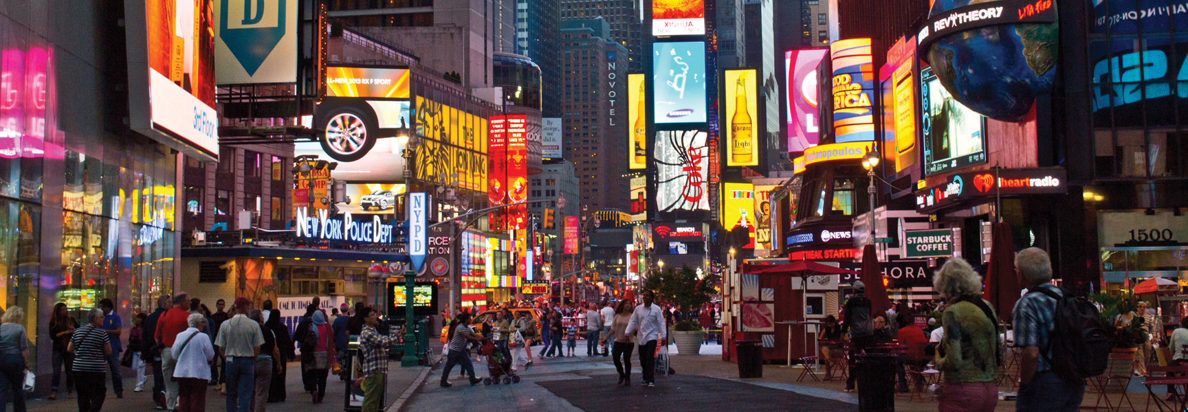 A panoramic view of New York's Times Square at dusk, with crowds of people walking below the iconic, brightly lit billboards.