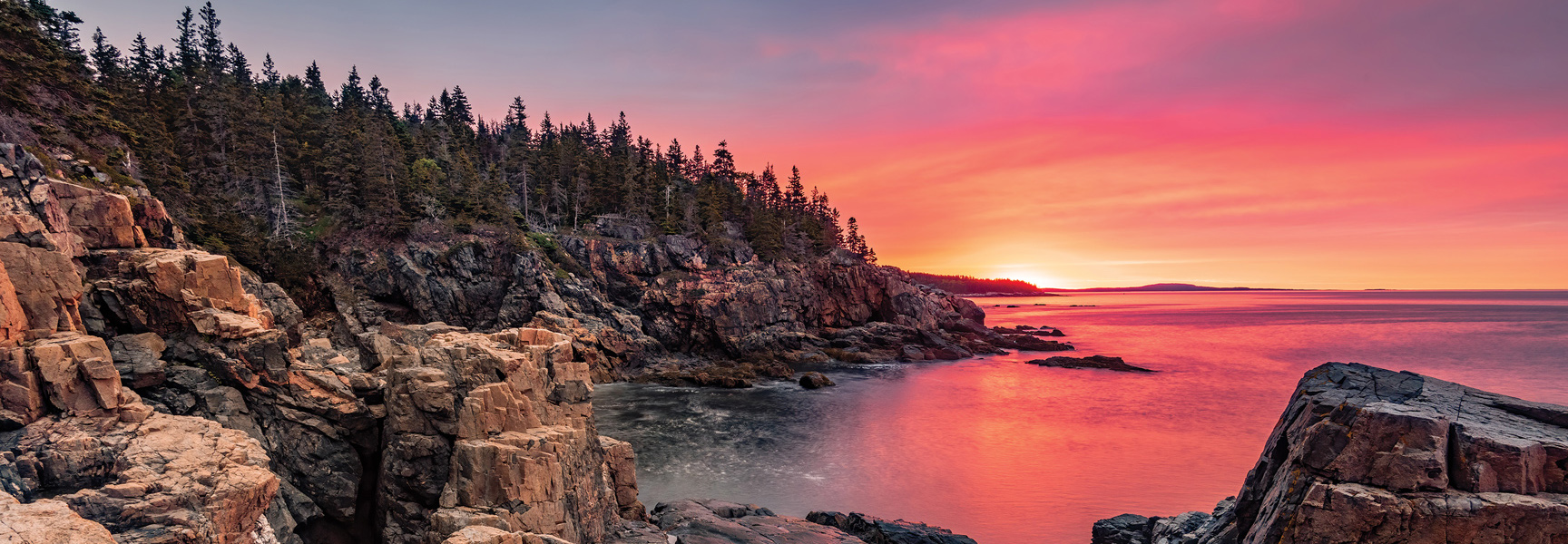 A vibrant pink and orange sunset glows over the rocky coastline of Acadia National Park in Maine.