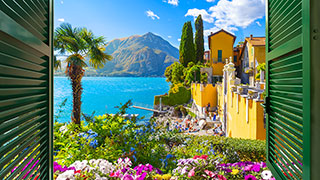 A view through green shutters reveals colorful villas and lush gardens along the shore of Lake Como, Italy, with mountains in the background.