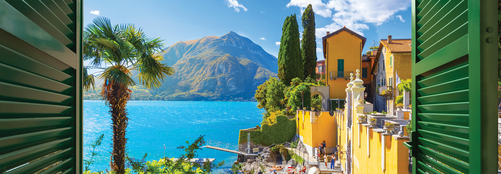 A view from a window with green shutters overlooks a sunny Italian lakeside village with colorful buildings, a bright blue lake, and large mountains.