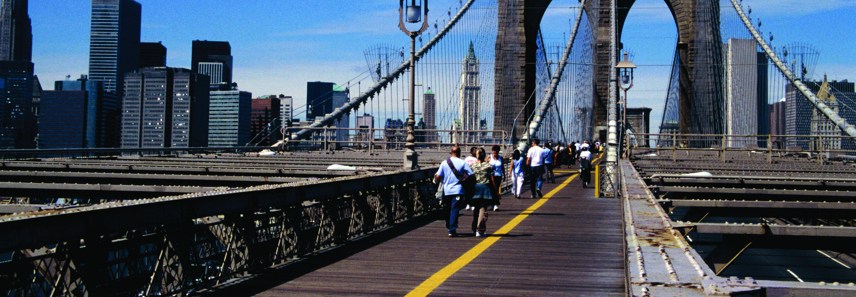 Pedestrians walk along the historic Brooklyn Bridge promenade towards the Manhattan skyline under a clear blue sky in New York.